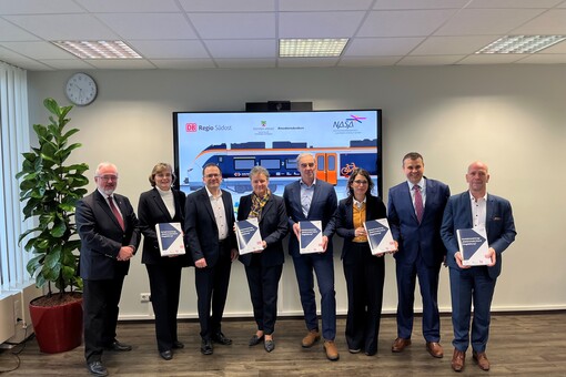 Drei Frauen und fünf Männer in dunklen Anzügen stehen in einem Raum vor einem Bildschirm. Auf dem Bildschirm ist das Modell eines orange-weißen Zuges zu sehen. Die Personen halten Vertragsunterlagen in den Händen.